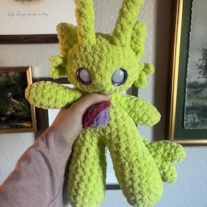 May include: A lime green crocheted creature plushie with large eyes and long antennae. The handmade toy has a small patch of pink and purple on its chest. The plushie is held up by a person.