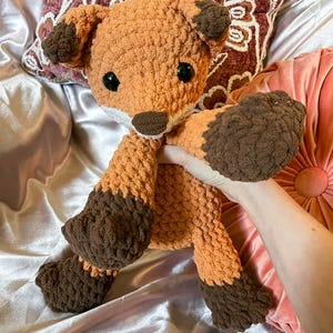 May include: A handmade crochet fox stuffed animal in shades of orange and brown. The fox has large black eyes, a white muzzle, and brown accents on its ears, paws, and feet. The plush toy is soft and cuddly, perfect for children or as a decorative item.
