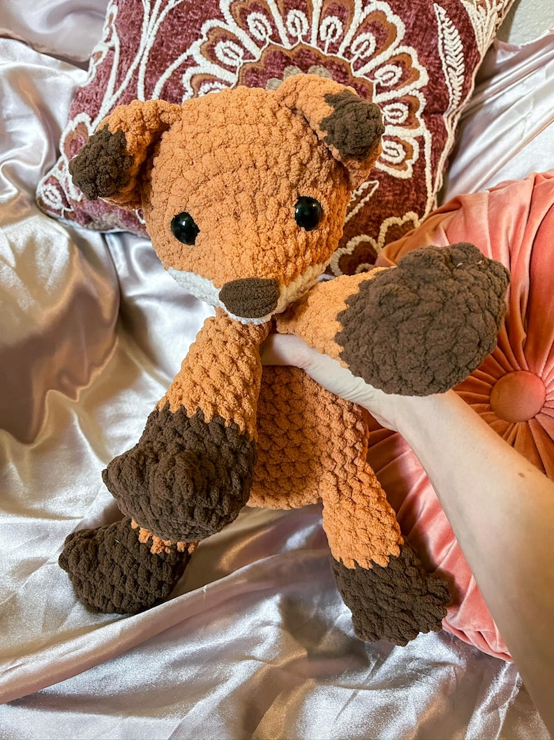 May include: A handmade crochet fox stuffed animal in shades of orange and brown. The fox has large black eyes, a white muzzle, and brown accents on its ears, paws, and feet. The plush toy is soft and cuddly, perfect for children or as a decorative item.