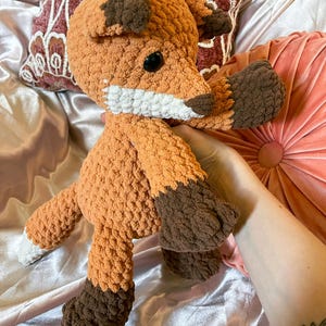 May include: Crocheted fox plush toy in shades of orange and brown. The fox has a white muzzle, black eyes, and brown accents on its ears and paws. The plush toy is soft and cuddly, perfect for children or as a decorative item.