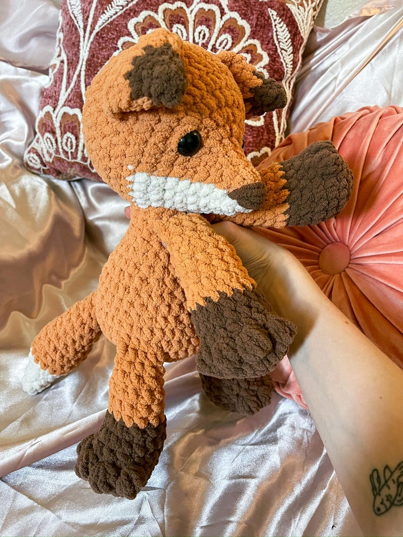 May include: Crocheted fox plush toy in shades of orange and brown. The fox has a white muzzle, black eyes, and brown accents on its ears and paws. The plush toy is soft and cuddly, perfect for children or as a decorative item.