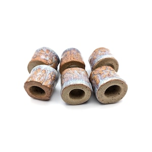 May include: A set of six cylindrical ceramic beads or connectors with a textured, earthy brown clay base and a glossy, crackled light blue and white glaze finish. Each piece features a hollow centre and a rugged, organic surface texture.