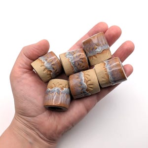 May include: Six small cylindrical ceramic beads with a tan base colour and a decorative carved sunburst pattern near the top. The upper portion of each bead features a glossy, dripped glaze in shades of blue, purple, and white over the tan clay.