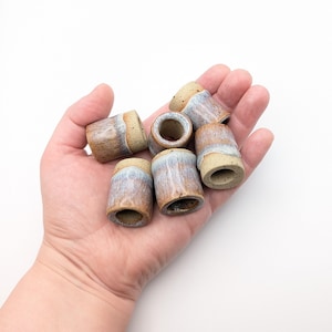 以下が含まれることがあります： 円筒形のハンドメイドセラミックビーズのコレクション。ビーズは、茶色、タン、青灰色の自然な色合いです。各ビーズの高さは約2.5cm、直径は約2cmです。