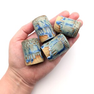 May include: Four small cylindrical ceramic cups featuring a textured, unglazed tan base and a reactive glaze finish in shades of blue, teal, and white that drips down the sides. Each cup has a distinct pattern of glaze pooling and colour variation.