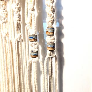 May include: A macrame wall hanging made of off-white cotton cord featuring intricate square knots and alternating vertical strands. The design incorporates cylindrical ceramic beads with a blue and tan glaze finish integrated into the knotted pattern.