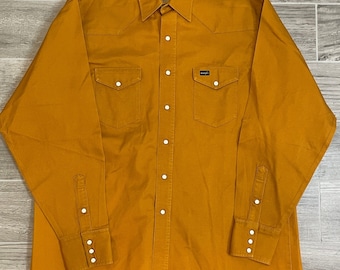 May include: A long-sleeved, button-down shirt in a mustard yellow color. The shirt has a pointed collar, two chest pockets with button closures, and a Wrangler brand label on the left chest.