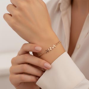 Op de afbeelding: Een roségoudkleurige kettingarmband met de naam ANNA in hoofdletters als centraal middelpunt. De armband bestaat uit een fijn schakelontwerp dat verbonden is met het naamplaatje, waardoor een doorlopende schakel om de pols ontstaat.