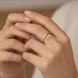 May include: A gold ring featuring a heart-shaped charm. The ring band is set with small, clear stones. The heart charm has a keyhole design. The ring is on a finger.