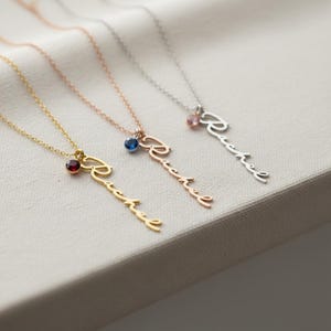 May include: Three necklaces featuring a delicate chain and a vertical pendant in cursive script reading Rachel. Each pendant is accented with a small round gemstone: a dark red stone on gold, a blue stone on rose gold, and a pink stone on silver.