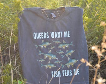 May include: Dark gray t-shirt with the text "QUEERS WANT ME FISH FEAR ME" in white. The shirt features an illustration of several fish in shades of blue and green.