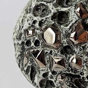 Puede incluir: Primer plano de una esfera de cerámica con una superficie texturizada. La esfera está cubierta de pequeños bultos en relieve y tiene un acabado plateado metálico. La superficie también está decorada con formas geométricas.