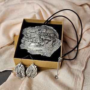 Puede incluir: Un conjunto de collar y pendientes de plata con un diseño texturizado y abstracto. El collar está en un cordón negro y los pendientes están sobre un fondo marrón.