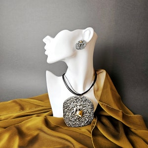 Puede incluir: Un collar de plata y oro con un gran colgante texturizado. El collar se muestra en un maniquí blanco con un par de pendientes de plata a juego.