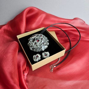 Puede incluir: Un conjunto de joyas que incluye un collar con colgante y pendientes a juego. El colgante y los pendientes tienen un acabado plateado texturizado con detalles rojos incrustados. El conjunto se presenta en una caja de cartón con interior negro.