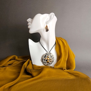 Puede incluir: Un maniquí de cerámica blanca que lleva un collar negro con un gran colgante redondo negro y dorado y un par de pendientes negros y dorados. El maniquí está drapeado en una tela amarilla mostaza.