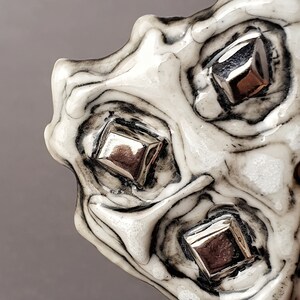 Puede incluir: Primer plano de un objeto decorativo con un patrón blanco y negro en espiral. El objeto tiene un acabado brillante y está adornado con tres adornos cuadrados de color plateado. El diseño es orgánico y abstracto.