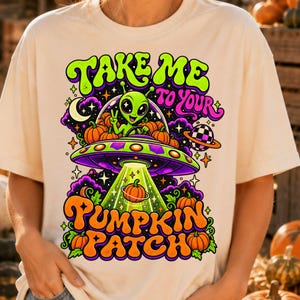 May include: A cream-colored t-shirt featuring a graphic of a green alien in a flying saucer surrounded by orange pumpkins. The text reads "TAKE ME TO YOUR PUMPKIN PATCH" in bold, colorful lettering. The design includes stars, a crescent moon, and a planet.