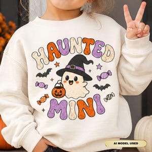 Puede incluir: Una sudadera de manga larga color crema con un gráfico de un fantasma sonriente que lleva un sombrero de bruja negro y sostiene un cubo de calabaza. El texto dice HAUNTED MINI, rodeado de ilustraciones de murciélagos, estrellas y caramelos.