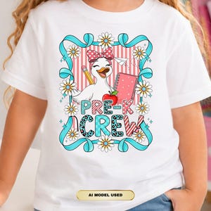 Può includere: Una t-shirt bianca con la grafica di un'anatra dei cartoni animati che indossa un fiocco rosso a pois, che tiene in mano una matita e una mela rossa accanto a un quaderno rosa. Il testo PRE-K CREW è stampato sotto, circondato da un nastro blu e margherite.