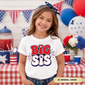 Könnte beinhalten: Ein weißes T-Shirt mit kurzen Ärmeln und dem Schriftzug BIG SIS in großen, fetten roten und weißen Buchstaben mit blauen Umrandungen. Das Design enthält kleine blaue Sterne und rot-blaue feuerwerksartige Grafiken.