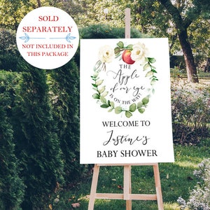 May include: A white sign with green and white floral wreath with a red apple in the center. The text reads "The Apple of our eye is on the way!"  Welcome to Justine's Baby Shower.