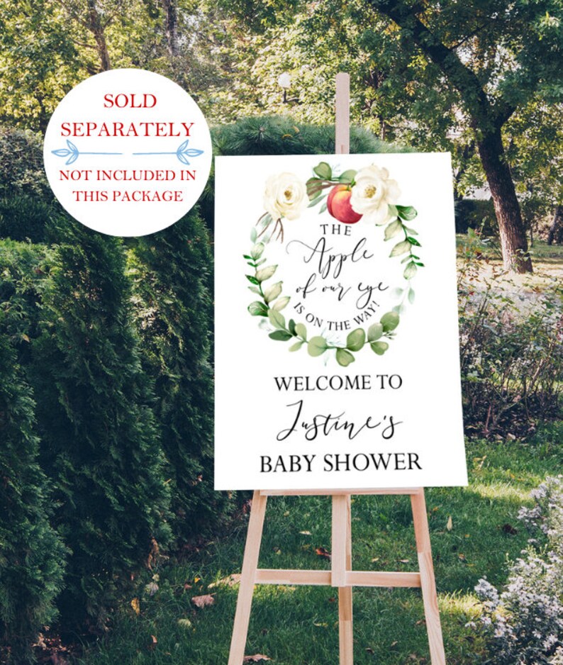 May include: A white sign with green and white floral wreath with a red apple in the center. The text reads "The Apple of our eye is on the way!"  Welcome to Justine's Baby Shower.