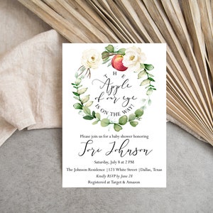 May include: A white baby shower invitation with a watercolor illustration of a red apple, white roses, and green leaves. The text reads "The Apple of our eye is on the way!" and "Please join us for a baby shower honoring [name] Saturday, July 8 at 2 PM The Johnson Residence 123 White Street Dallas, Texas Kindly RSVP by June 28 Registered at Target & Amazon"