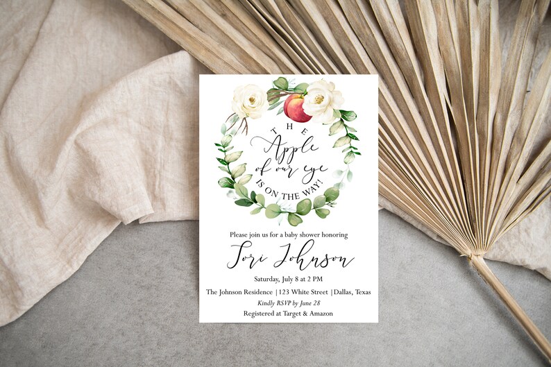 May include: A white baby shower invitation with a watercolor illustration of a red apple, white roses, and green leaves. The text reads "The Apple of our eye is on the way!" and "Please join us for a baby shower honoring [name] Saturday, July 8 at 2 PM The Johnson Residence 123 White Street Dallas, Texas Kindly RSVP by June 28 Registered at Target & Amazon"
