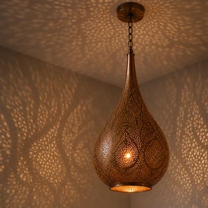 Peut inclure: Un luminaire suspendu en bronze en forme de goutte d'eau. L'abat-jour en métal est finement perforé, projetant une lumière à motifs sur les murs et le plafond environnants. Le luminaire est suspendu à une chaîne fixée à un support de plafond circulaire.