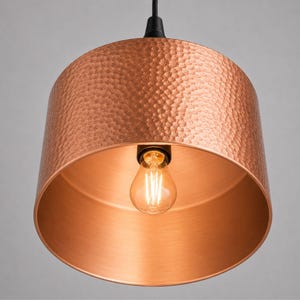 May include: A copper pendant light fixture with a hammered texture on the exterior. The interior is smooth and reflective, with a visible lightbulb. The fixture hangs from a black cord and canopy, creating a warm, inviting ambiance. The light fixture is a decorative element.