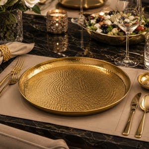 Peut inclure: Une assiette ronde en métal doré avec une texture martelée et alvéolée sur toute sa surface. L'assiette présente un bord légèrement surélevé et une finition polie, reflétant la lumière sur son motif circulaire texturé sur un set de table neutre.