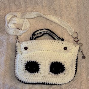 May include: A handmade, white crochet bag with a black trim and two large, black circular accents. The bag features a white strap, a small handle, and a decorative charm. Two brown buttons secure the flap.