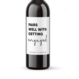 May include: A white wine bottle label with black text that reads "Pairs well with getting engaged. Cheers to forever."