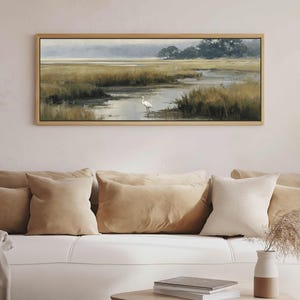 May include: A rectangular landscape painting in a light wood frame depicting a marshland scene with tall golden grasses, a winding waterway, and a single white egret standing in the water under a soft, muted sky with distant dark trees.