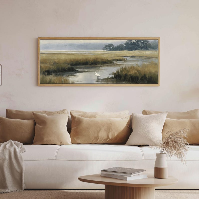 May include: A rectangular landscape painting in a light wood frame depicting a marshland scene with tall golden grasses, a winding waterway, and a single white egret standing in the water under a soft, muted sky with distant dark trees.