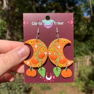May include: A pair of orange crescent moon earrings with dangling pumpkins and green leaves. The earrings are decorated with white stars, yellow flowers, and green vines. The earrings are on a brown card with the text "Clafful Tinker".