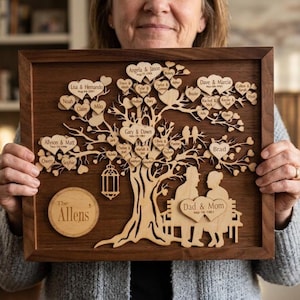 Può includere: Un'opera d'arte ad albero genealogico in legno con una cornice marrone scuro. L'albero presenta foglie a forma di cuore con nomi, una panchina con silhouette e le parole "Dad & Mom". Viene visualizzato anche il cognome "The Allens".