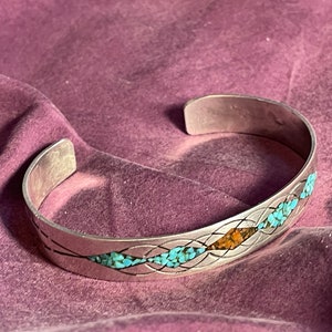 Op de afbeelding: Een zilveren armband met een geweven ontwerp en ingelegde turquoise en oranje stenen.