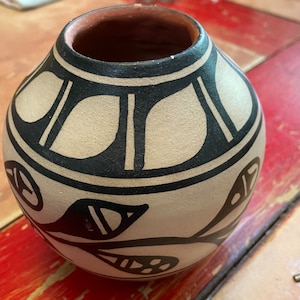 Op de afbeelding: Een kleine, ronde, beige aardewerken vaas met zwarte geverfde ontwerpen. De vaas heeft een geometrisch patroon rond de bovenkant en een bloemmotief aan de onderkant.