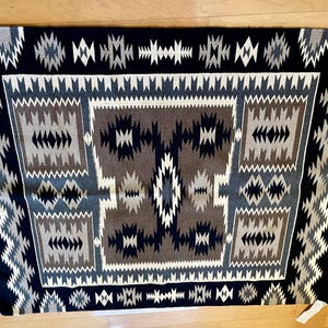 Może przedstawiać: Navajo dywan z geometrycznym wzorem w odcieniach czerni, brązu, bieli i szarości. Dywan ma centralny kwadrat z mniejszym kwadratem wewnątrz, otoczony obramowaniem z powtarzających się geometrycznych kształtów.