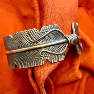 May include: A silver-toned bracelet with a detailed feather design. The bracelet is made of metal and has a textured finish.