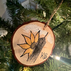 May include: A hand-painted wooden ornament featuring a stylized sunflower and a skyscraper with white clouds in the background. The ornament is decorated with gold paint and has the initials "KB" written on the bottom.