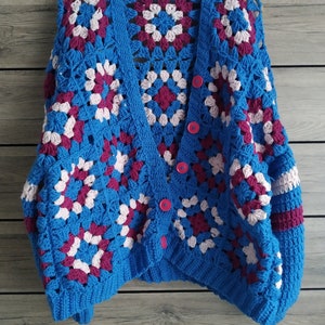 Puede incluir: Un cárdigan de crochet azul con detalles en rosa y burdeos. El cárdigan tiene cierre de botón y un ajuste holgado.