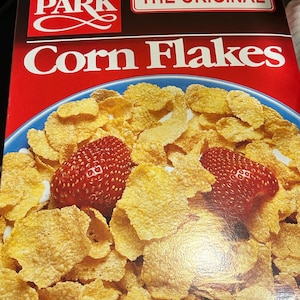 Könnte beinhalten: Eine Packung Hyde Park The Original Corn Flakes Müsli. Die Packung hat einen roten Hintergrund mit weißem Text und ein Bild von Cornflakes mit Erdbeeren darauf. Auf der Packung steht "Nettogewicht 18 oz (1 lb 2 oz) 510 Gramm."