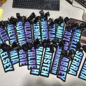 Könnte beinhalten: Eine Sammlung rechteckiger Glitzer-Schlüsselanhänger mit blau-violetten Farbverlaufshintergründen und schwarzen Namenstexten wie Allie, Jaelyn, Sophia, Khloe, Hannah, Kirsten, Carrington, Abbey, Sierra und anderen, jeweils mit einer schwarzen Schleife.