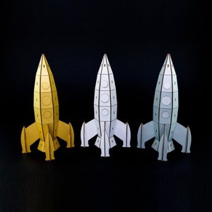 May include: Three paper rockets in different colors: yellow, white, and light blue. The rockets are made of cardboard and have a simple design. They are all facing forward and have a pointed nose cone.