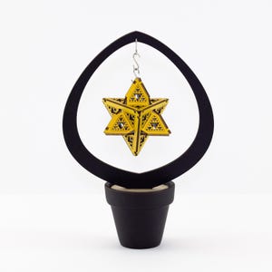 May include: A gold geometric star-shaped ornament hangs from a black wooden stand. The stand has a pot-like base.
