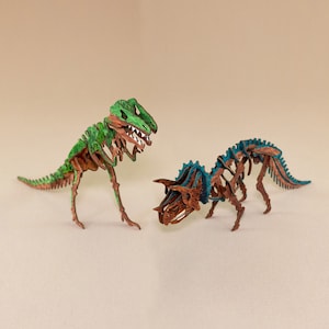 May include: Two wooden dinosaur models, one green and one blue, with intricate details and a skeletal design. The green dinosaur is a Tyrannosaurus Rex, while the blue dinosaur is a Triceratops.