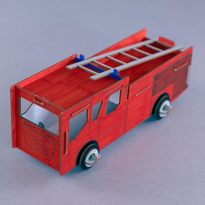 May include: A red wooden toy fire engine with a ladder on top. The toy has black wheels and white accents. The text "FIRE" is visible on the side of the vehicle.